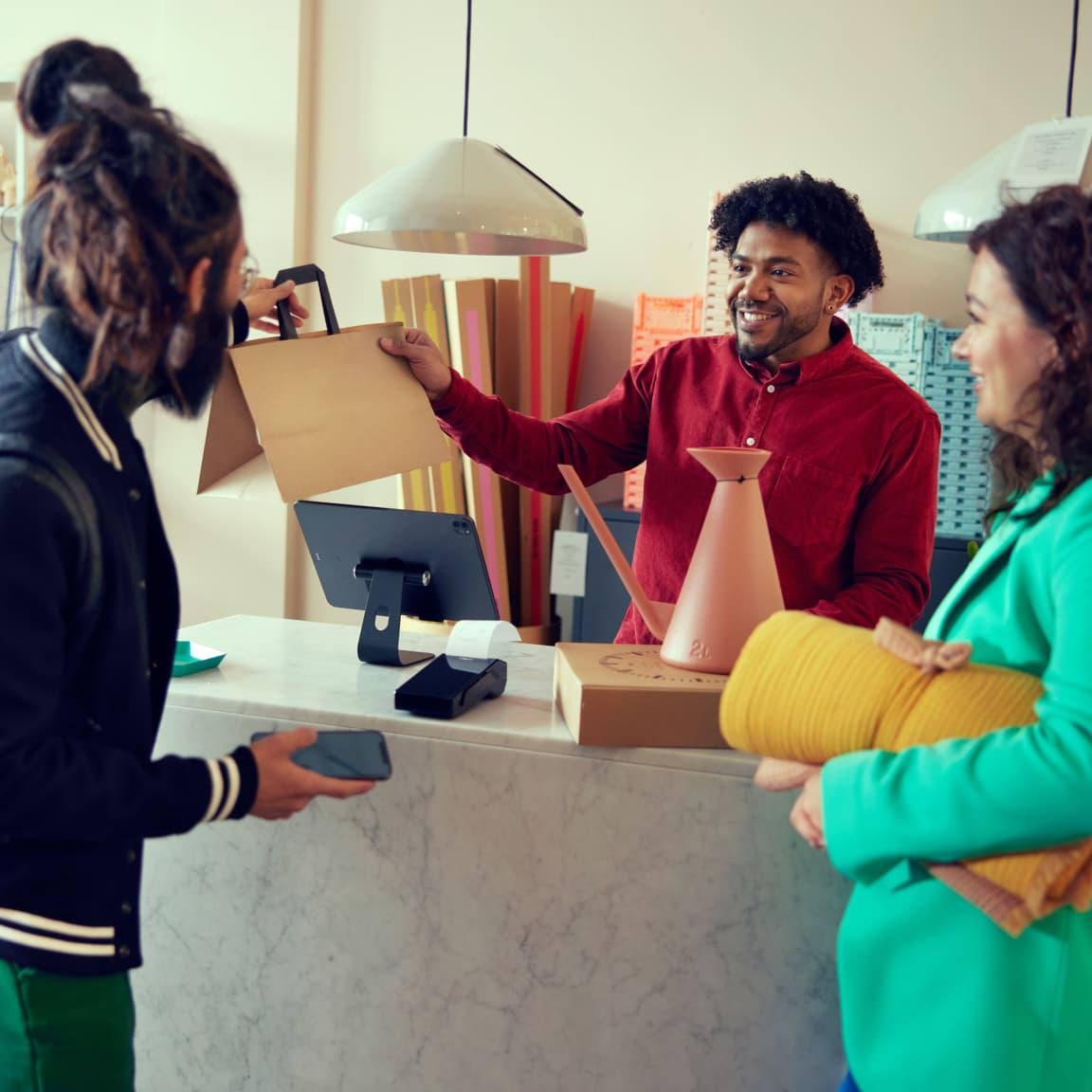 A smiling cashier handing over a product to a customer with a point of sale terminal on the counter.